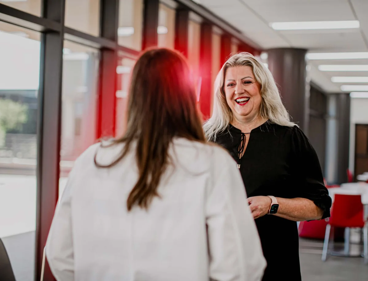 Faculty member speaking with a healthcare student in a campus hallway near floor-to-ceiling windows
