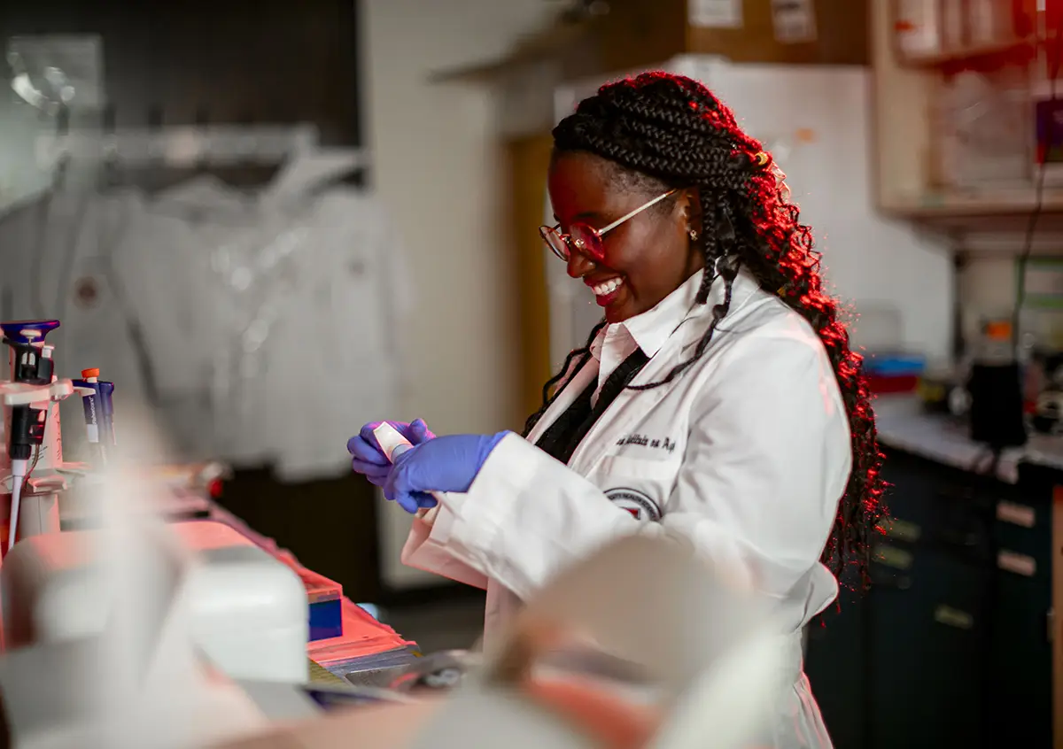 Student in white lab coat and gloves handling a sample in a biomedical research laboratory