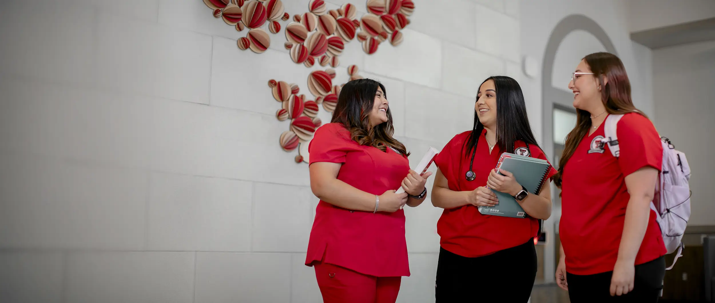 three students in red scrubs talking together while holding books